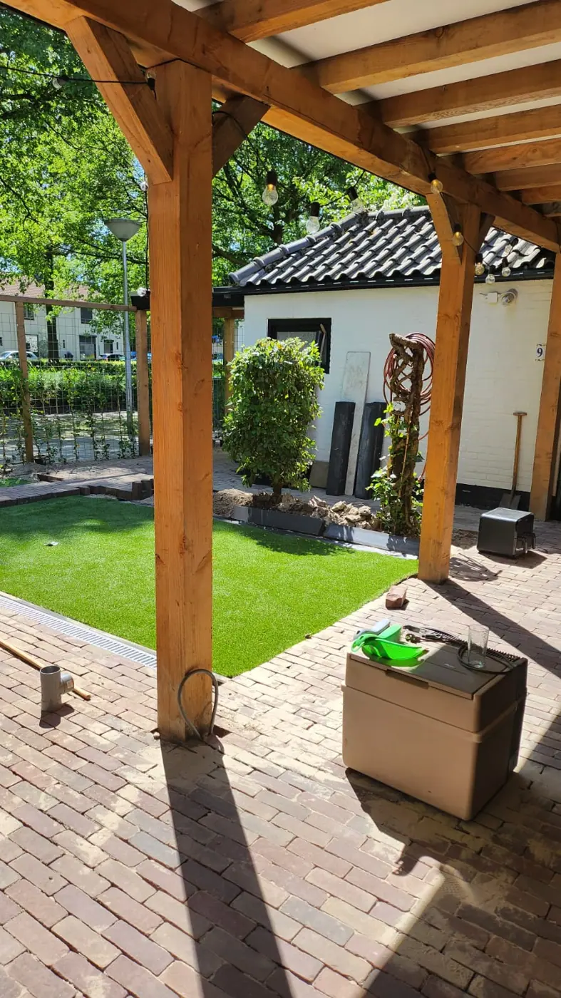 Houten staanders van een overkapping naast kunstgras en klinkers
