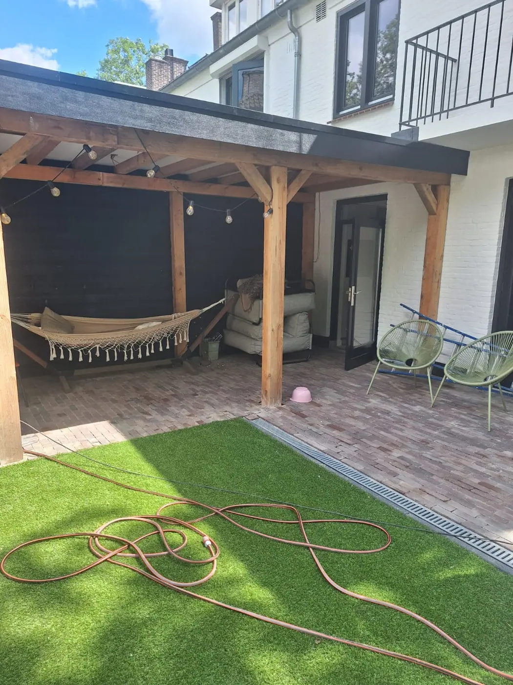 Houten overkapping aan het huis met terras, kunstgras en een hangmat